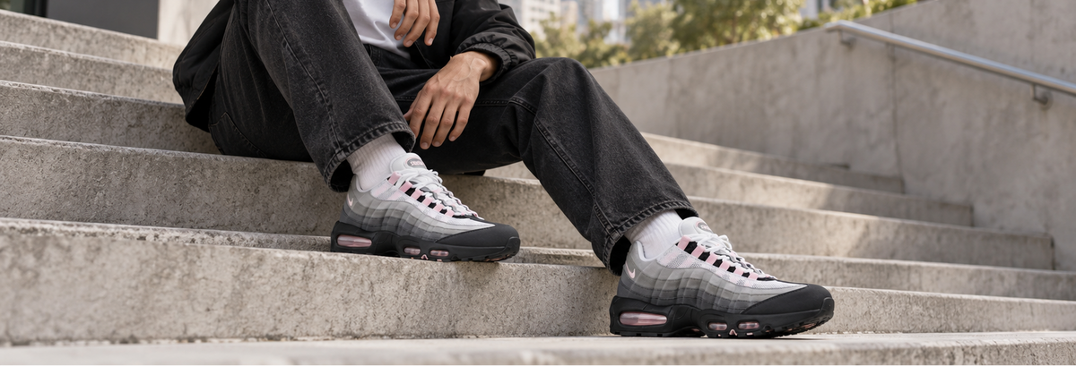 A person sitting on concrete outdoor steps wearing a pair of low-top athletic sneakers with a gray gradient upper, black sole, white laces, and pink accents.